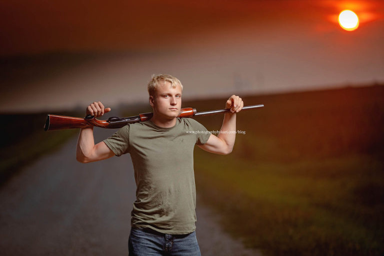 senior boy with wiuth gun over shoulders on gravel with road with sun setting behind