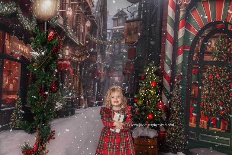 christmas mini photo of little girl wearing red and green plaid dress holding a present taken in studio by photography by kari