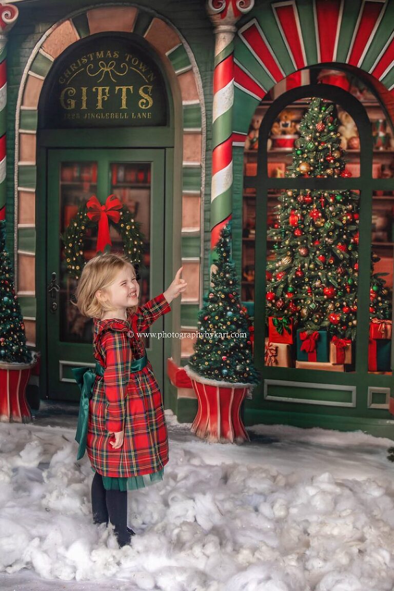 christmas mini photo of little girl wearing red and green plaid dress pointing at a toy throw a small store window taken in studio by photography by kari